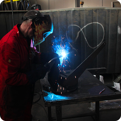Welder fabricating a steel marine (Knox) anchor component in a workshop.