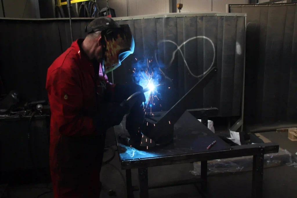 Welder fabricating a steel marine (Knox) anchor component in a workshop.